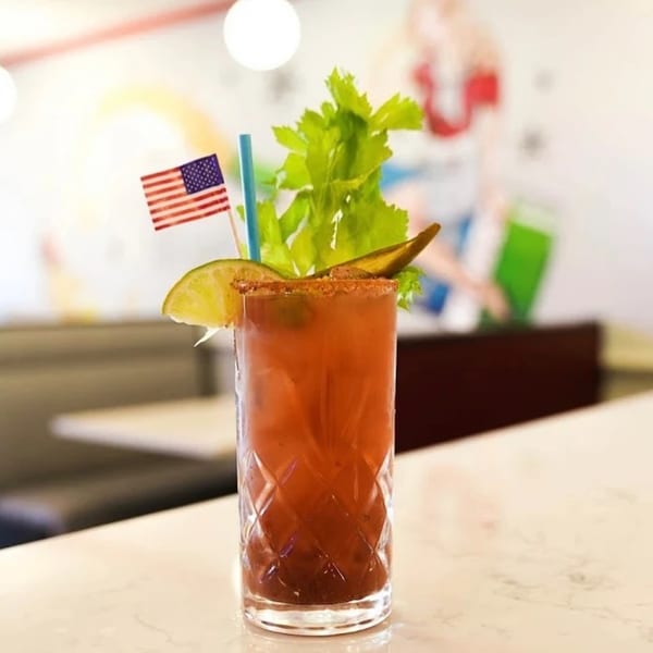 A Bloody Mary cocktail with celery, lime, and an American flag pick on a white bar.