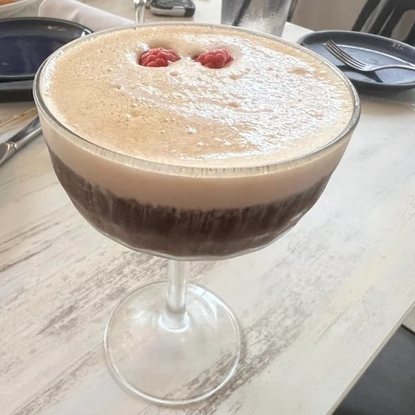 A dark cocktail with a thick layer of foam and two raspberries on top, on a white table.