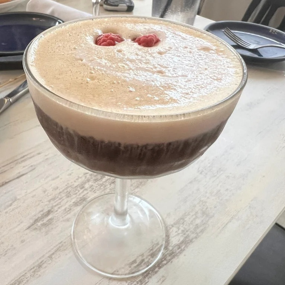 A dark cocktail with a thick layer of foam and two raspberries on top, on a white table.