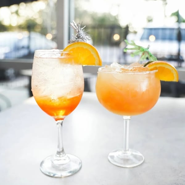 Two orange cocktails with ice and orange slices, one with a silver tinsel pick, on a table.