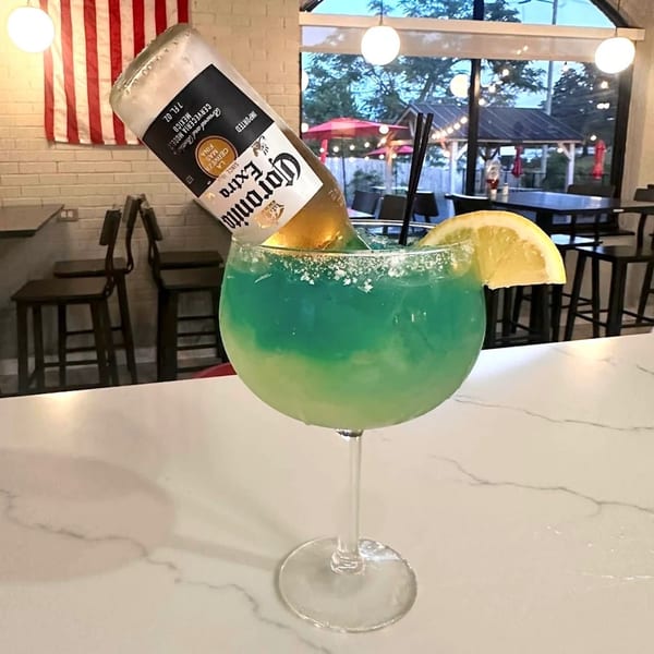 A large blue-green margarita with a Corona beer bottle inverted into it, garnished with a lemon slice.