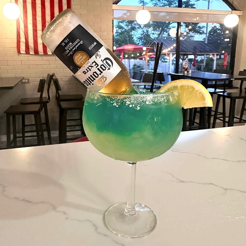 A large blue-green margarita with a Corona beer bottle inverted into it, garnished with a lemon slice.